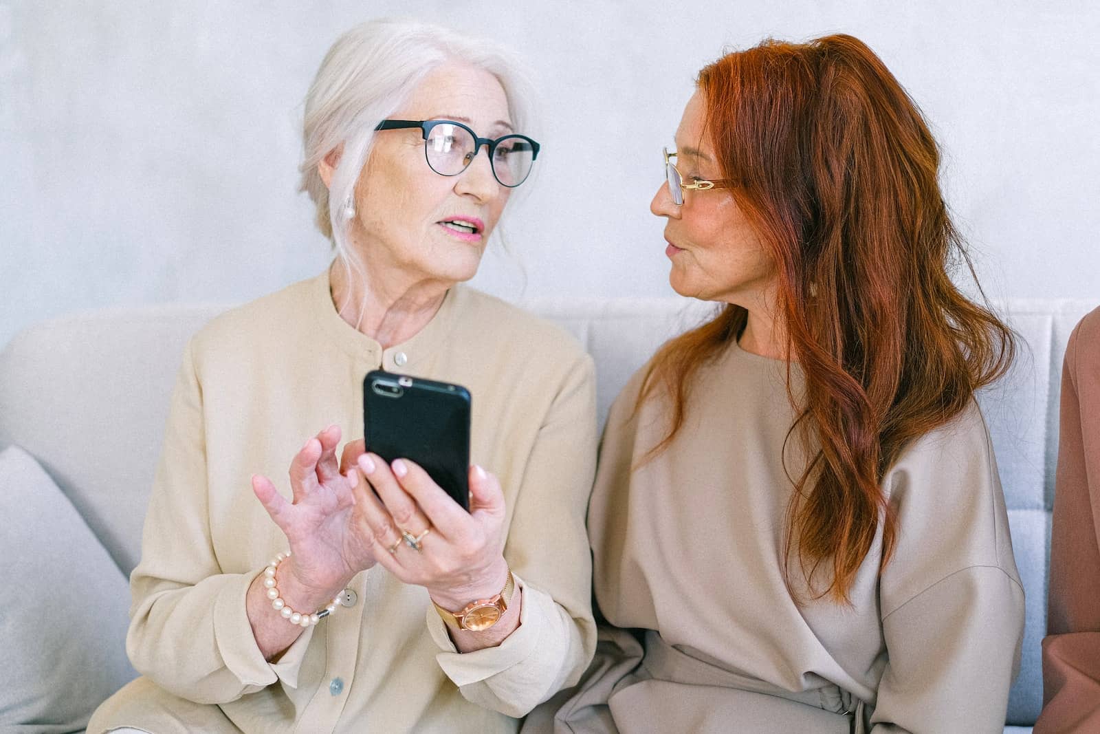 Elderly women in casual clothes sitting on sofa and communicating while browsing smartphone in light room